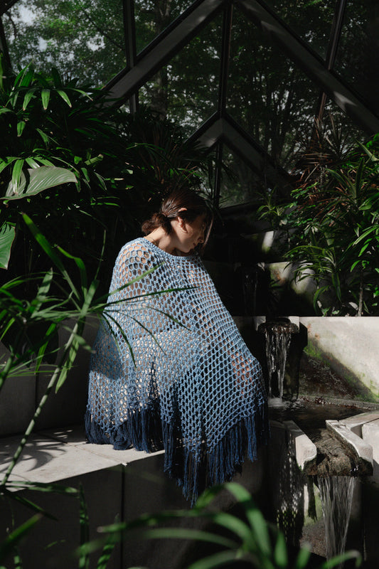 Woman wrapped in a blue crochet shawl sitting by a fountain with greenery around