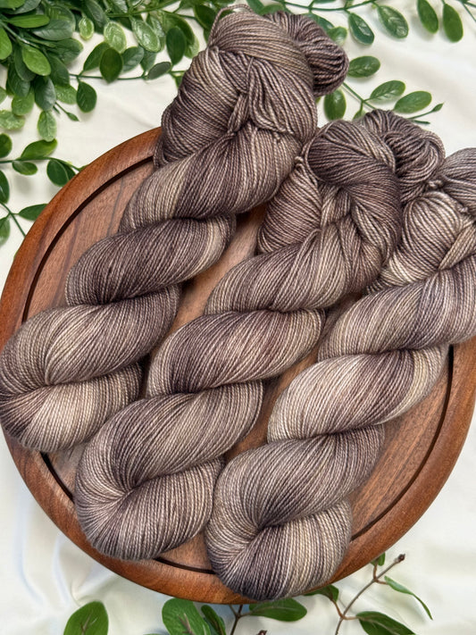Three hanks of variegated yarn in a dusty mauve-brown and taupe colorway, arranged on a round wooden board with green leaves on a white fabric background.