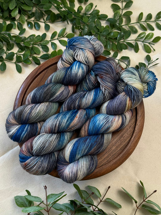 Three skeins of variegated yarn with blue, gray, and cream stripes, arranged on a round wooden tray with green leaves around it.