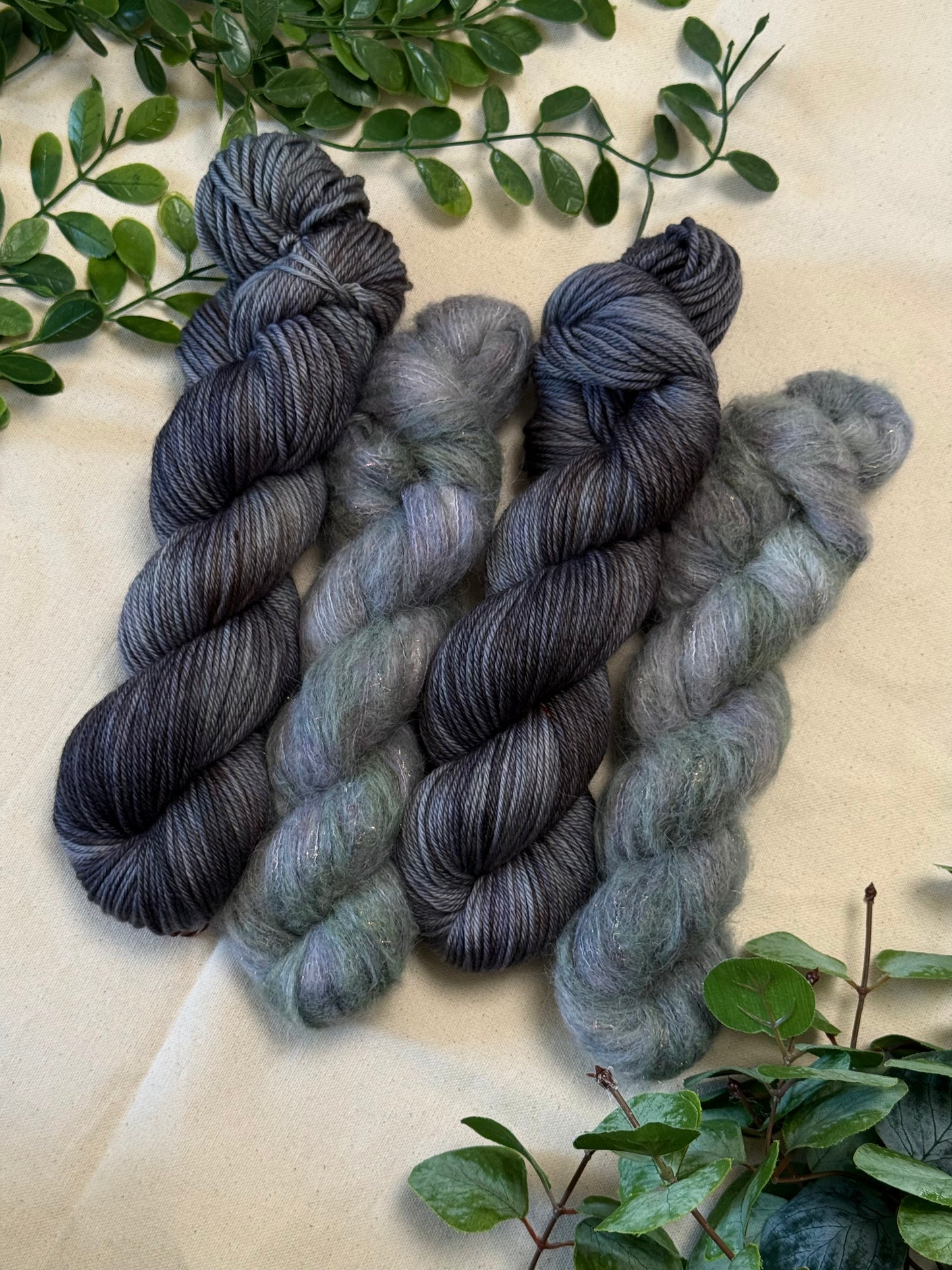Four hanks of variegated yarn arranged on a beige fabric background with green foliage accents. The yarn transitions through dark charcoal, black, and slate gray into muted sage green and light gray.
