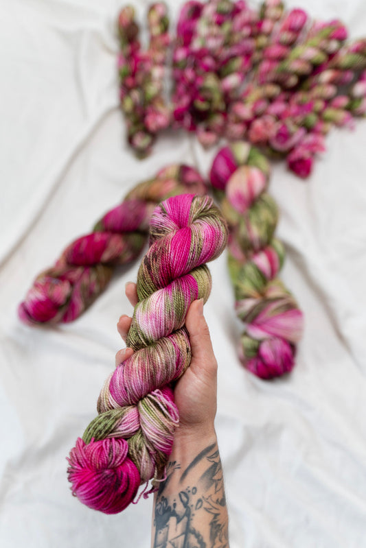 Hand holding a skein of pink and green yarn with more yarn skeins in the background on a white surface.