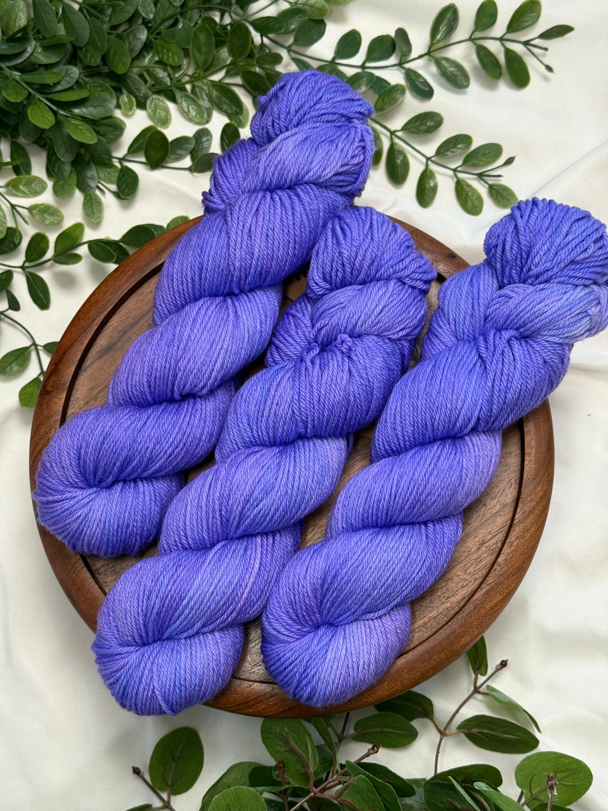 Three skeins of periwinkle blue-violet yarn arranged on a round wooden tray, surrounded by green foliage on a white fabric background.