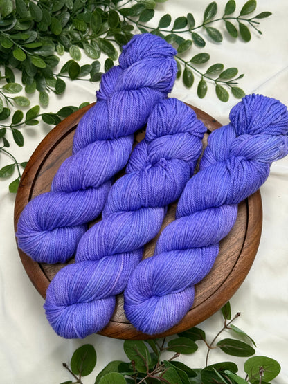 Three skeins of periwinkle blue-violet yarn arranged on a round wooden tray, surrounded by green foliage on a white fabric background.