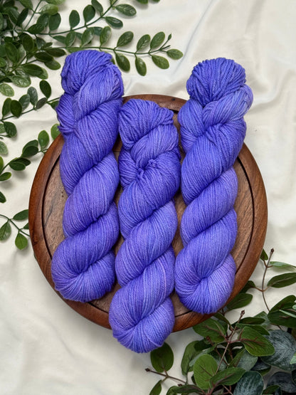 Three skeins of periwinkle blue yarn arranged on a round wooden tray, surrounded by green leaves on a white fabric background.