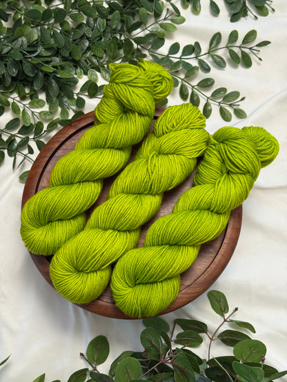 Three bright lime green skeins of yarn arranged on a round wooden tray, surrounded by green leaves on a white fabric background.