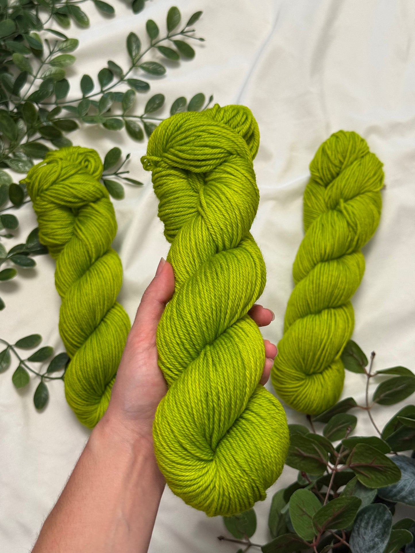 Three skeins of bright chartreuse green yarn arranged on a white fabric background with green leaves. A hand holds the center skein.