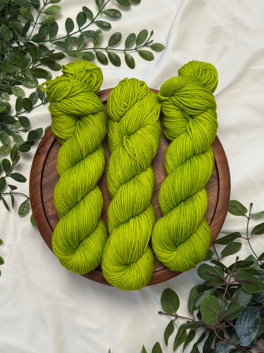 Three bright lime green skeins of yarn arranged on a round wooden board, styled with green leaves on a white fabric background.