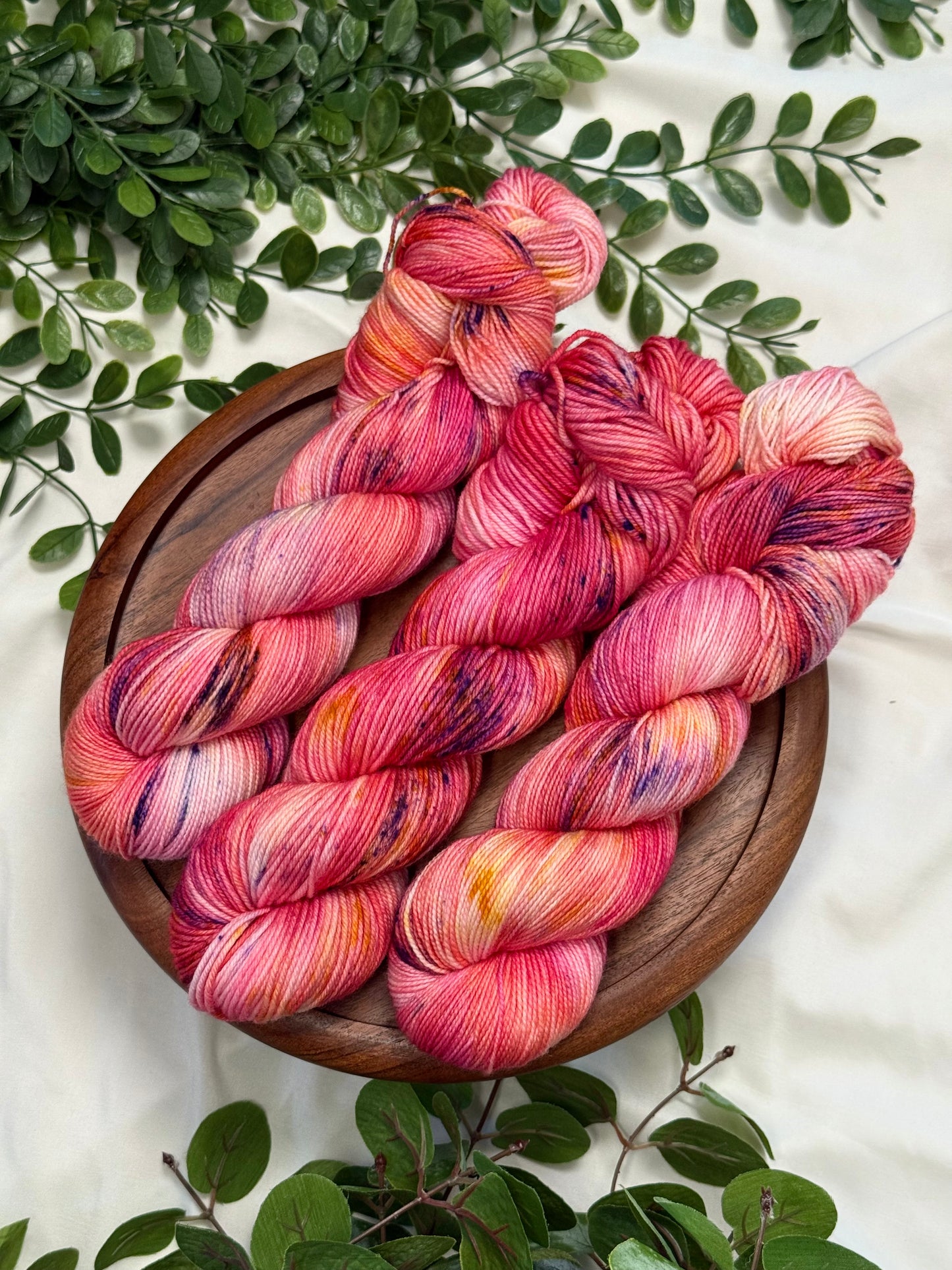 Four hanks of variegated yarn in shades of pink, red, orange, and purple, arranged on a round wooden board with green leaves on a white fabric background.