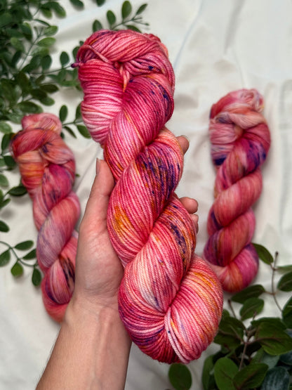 A hand holds a twisted hank of variegated pink and orange yarn with purple flecks, with two more skeins placed on a white fabric background decorated with green leaves.
