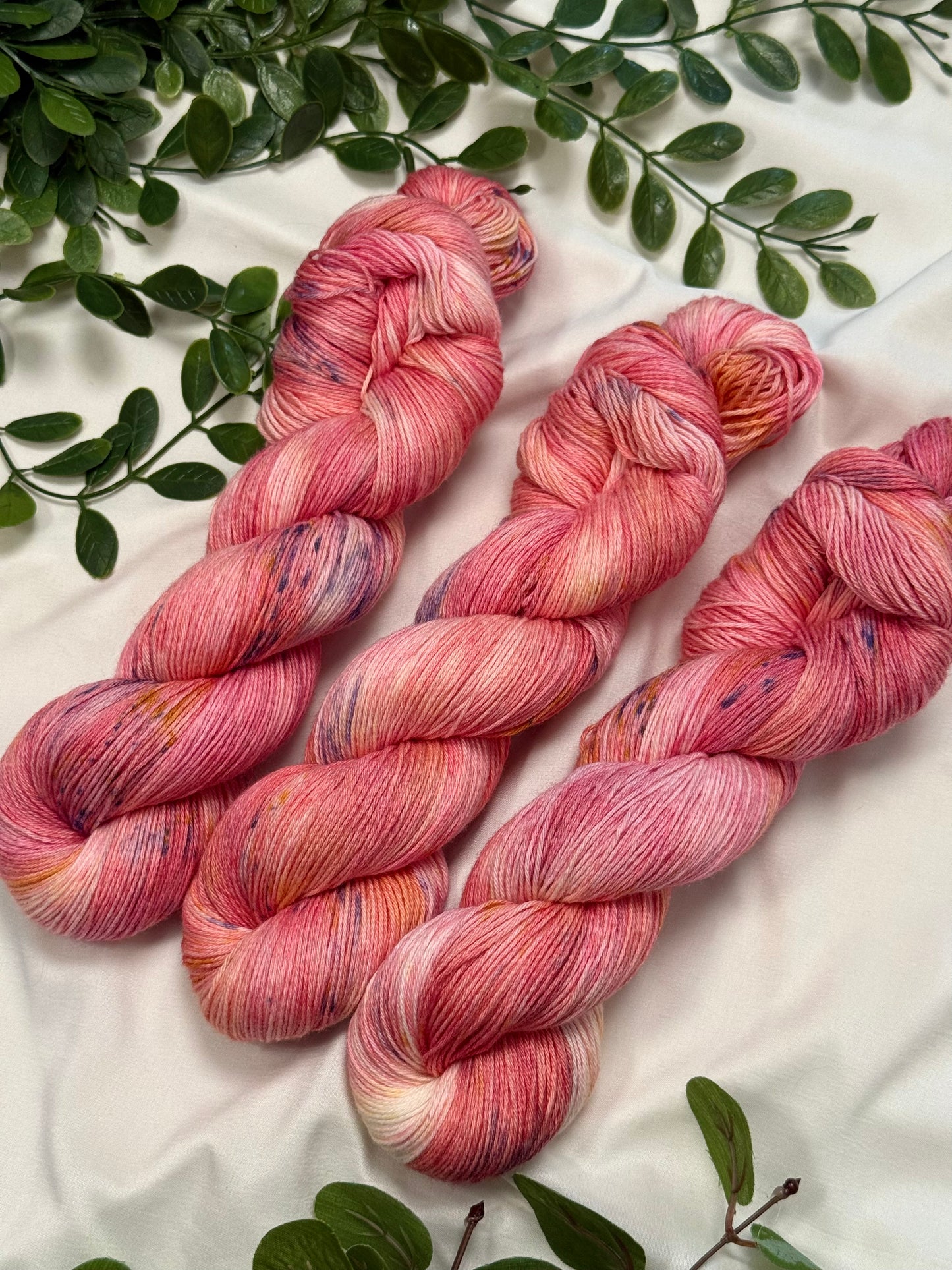 Three twisted hanks of variegated pink and peach yarn with subtle purple and cream flecks, arranged on a white fabric background with green leaves.