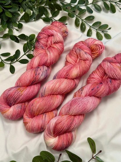 Three twisted hanks of variegated pink and peach yarn with subtle purple and cream flecks, arranged on a white fabric background with green leaves.