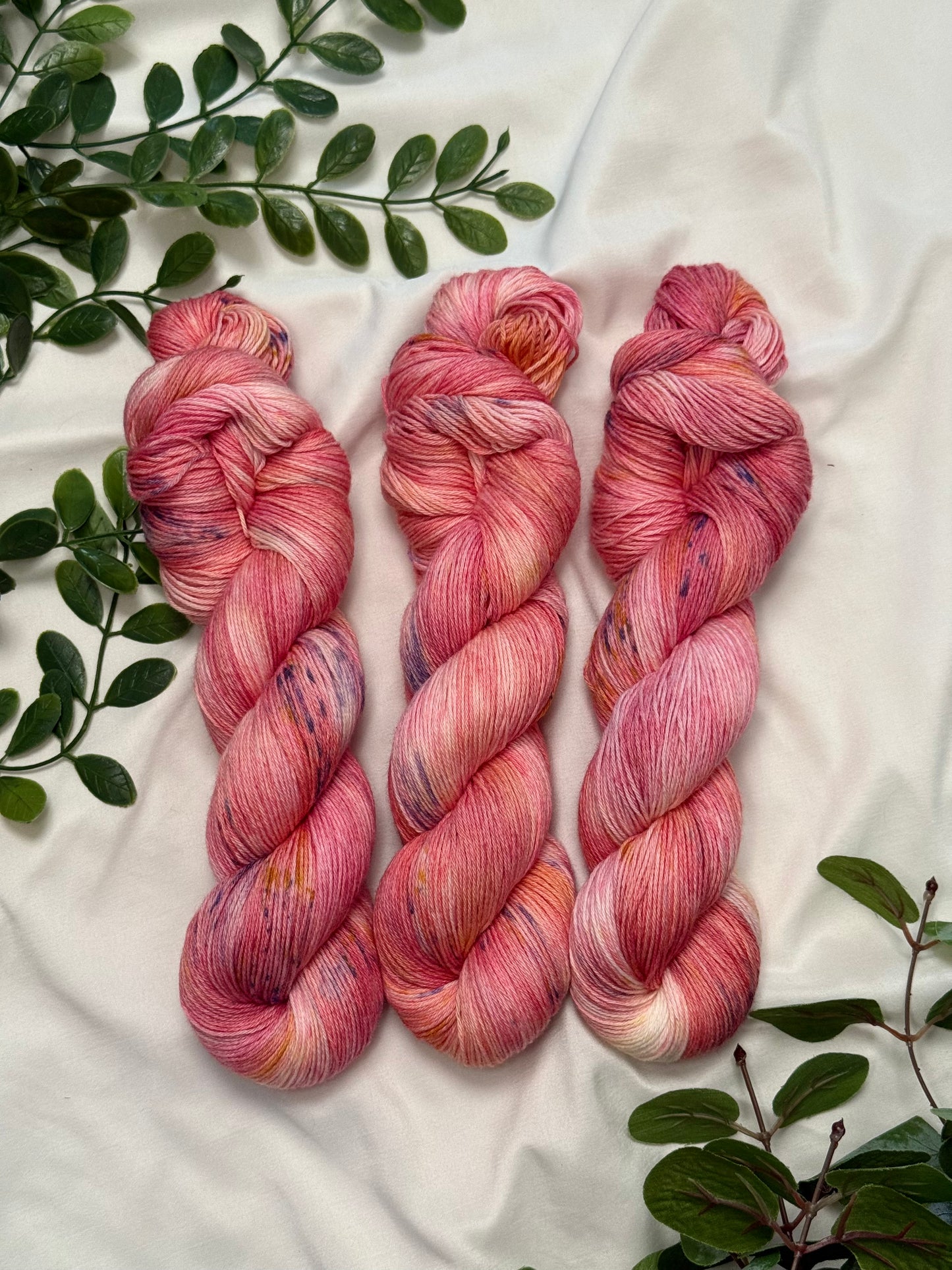 Three twisted hanks of variegated pink and peach yarn with purple and yellow flecks, arranged on a white fabric background with green leaves.