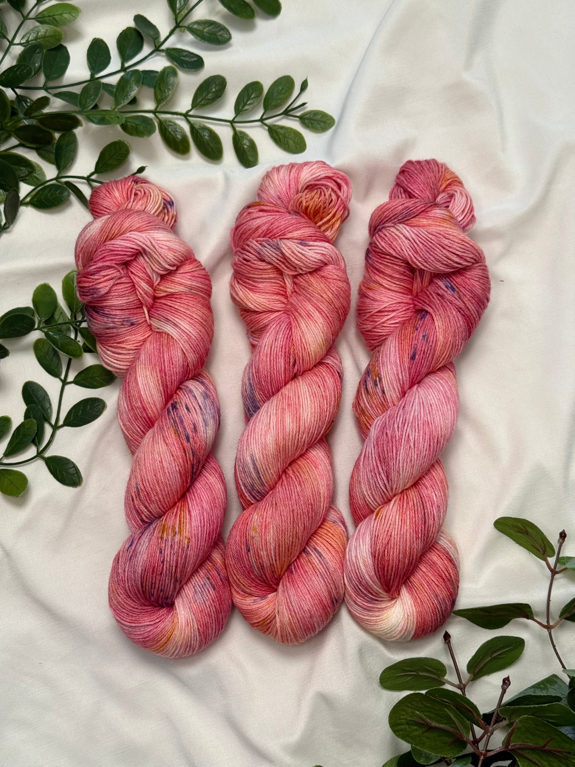 Three twisted hanks of variegated pink and peach yarn with purple and yellow flecks, arranged on a white fabric background with green leaves.
