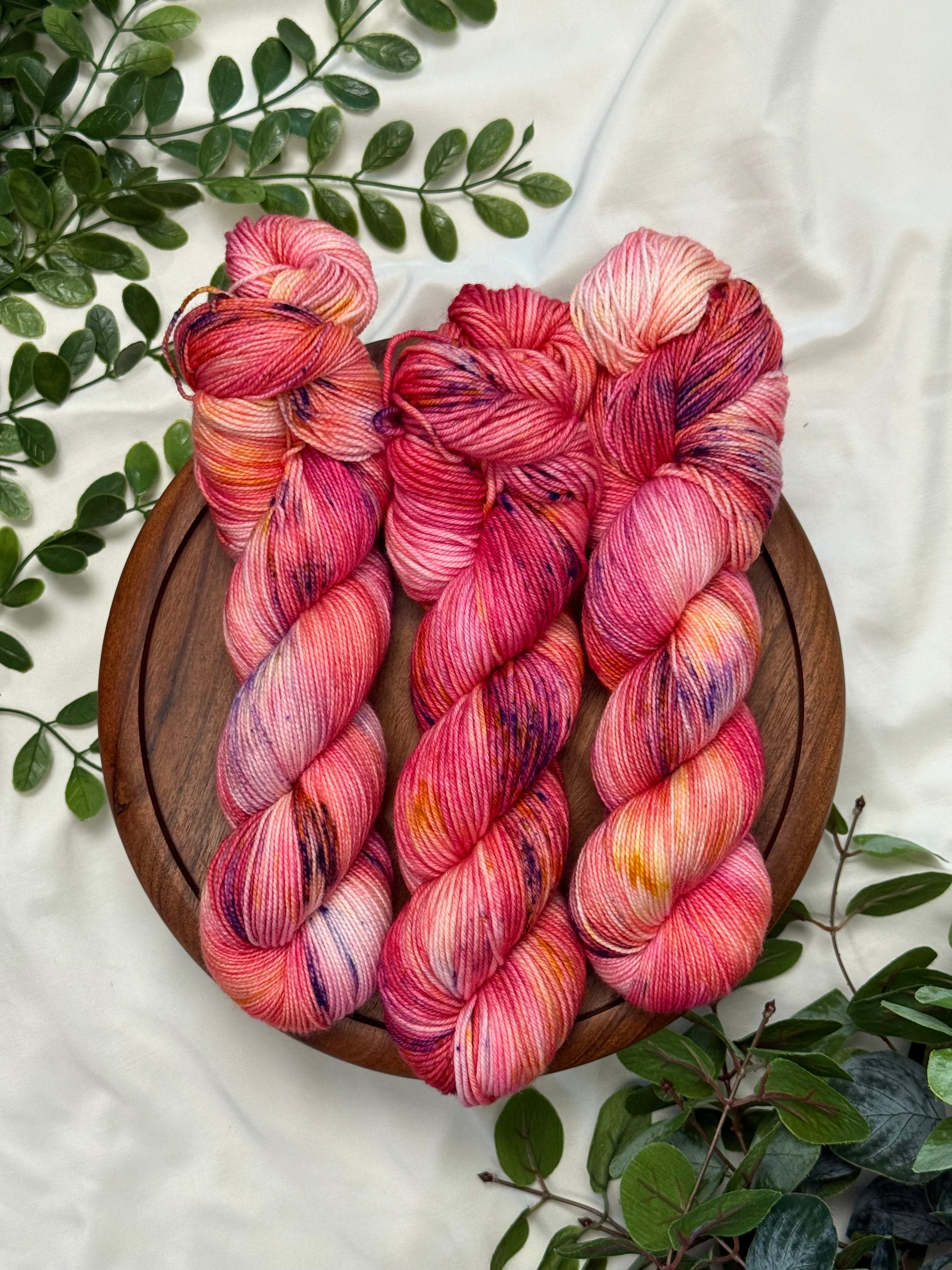 Three twisted hanks of variegated pink and red yarn with purple and orange flecks, arranged on a round wooden tray with green leaves on a white fabric background.