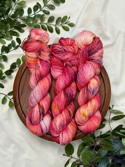 Three twisted hanks of variegated pink and red yarn with purple and orange flecks, arranged on a round wooden tray with green leaves on a white fabric background.