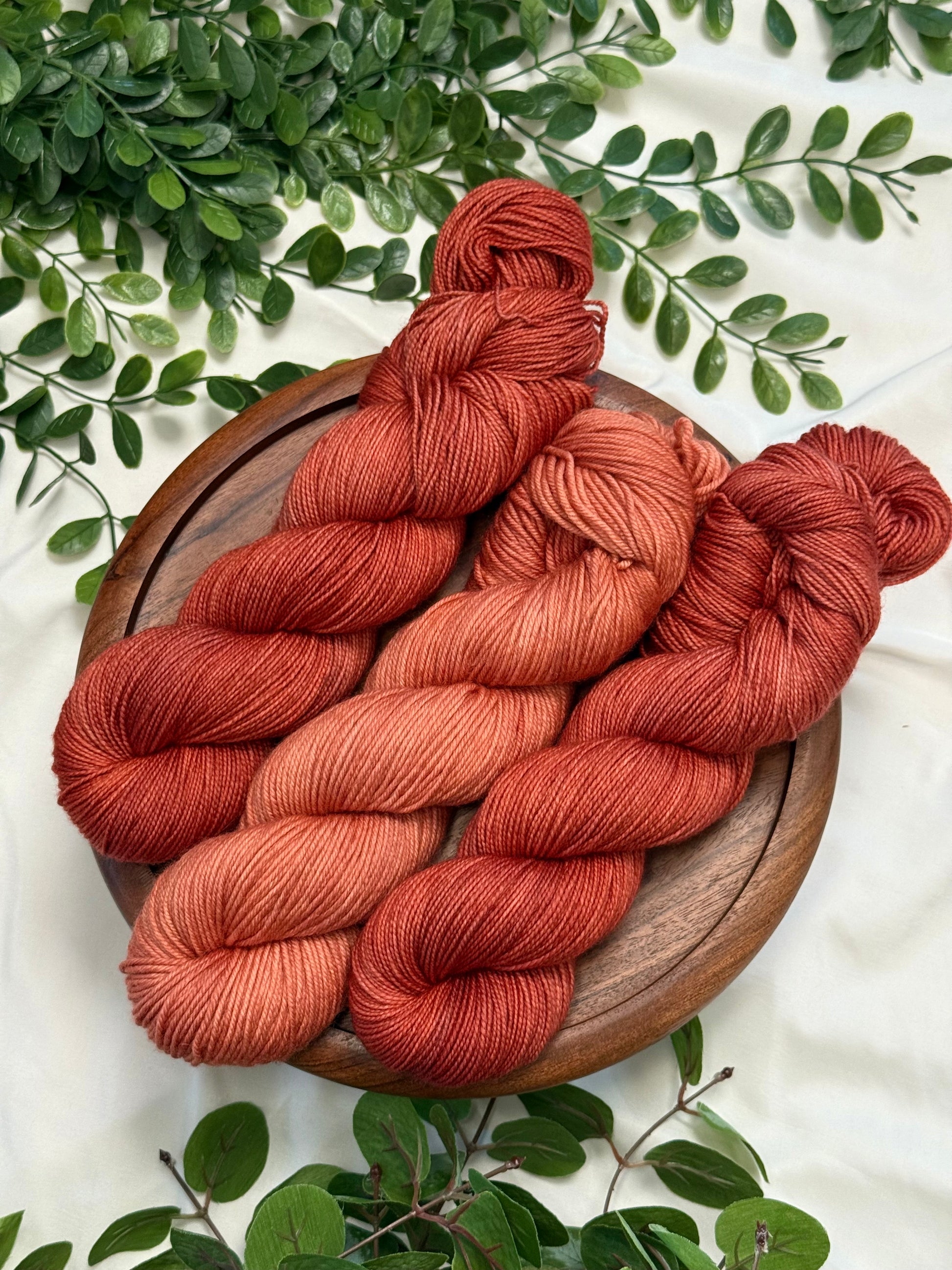 Three skeins of twisted yarn in warm rust, terracotta, and burnt orange shades, arranged on a round wooden tray with green foliage around it on a white fabric background.