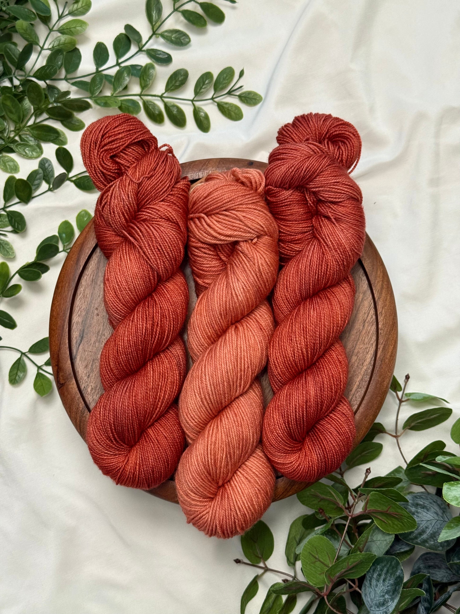 Three twisted skeins of yarn in warm rust, terracotta, and burnt orange shades, arranged on a round wooden board with green leaves on a light fabric background.