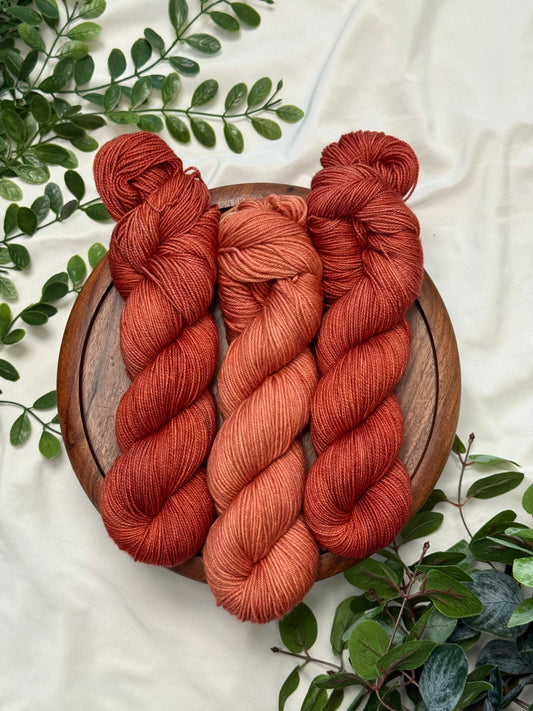 Three twisted skeins of yarn in warm rust, terracotta, and burnt orange shades, arranged on a round wooden board with green leaves on a light fabric background.