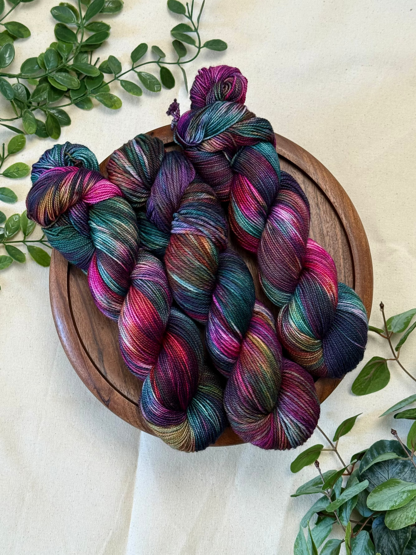 Four skeins of variegated yarn with a rich blend of teal, purple, magenta, and brown tones, arranged on a round wooden tray and decorated with green leaves on a cream fabric background.
