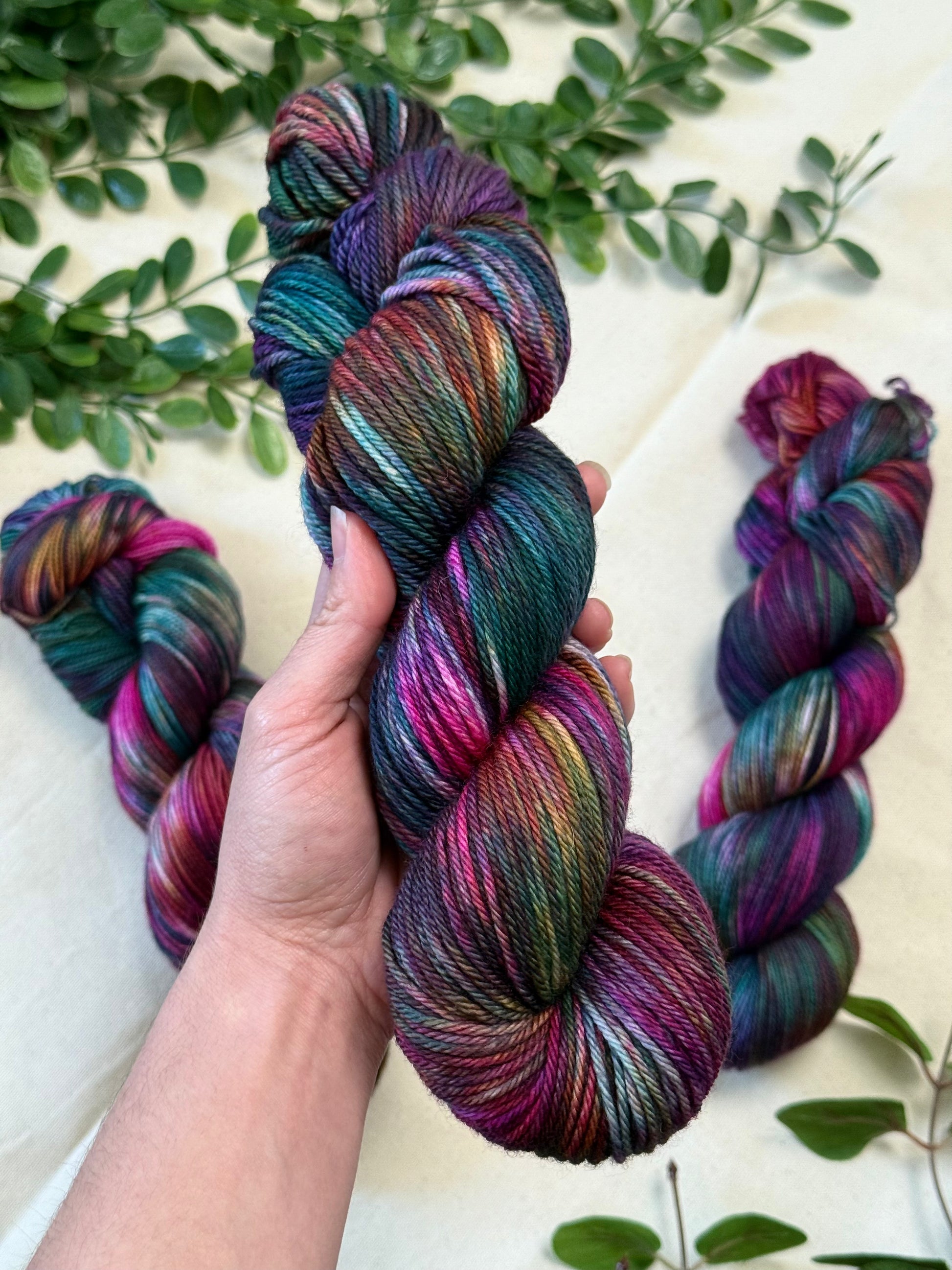 A hand holds a twisted skein of variegated yarn with rich purple, teal, green, and pink tones, with three more skeins arranged on a light fabric background with green leaves.