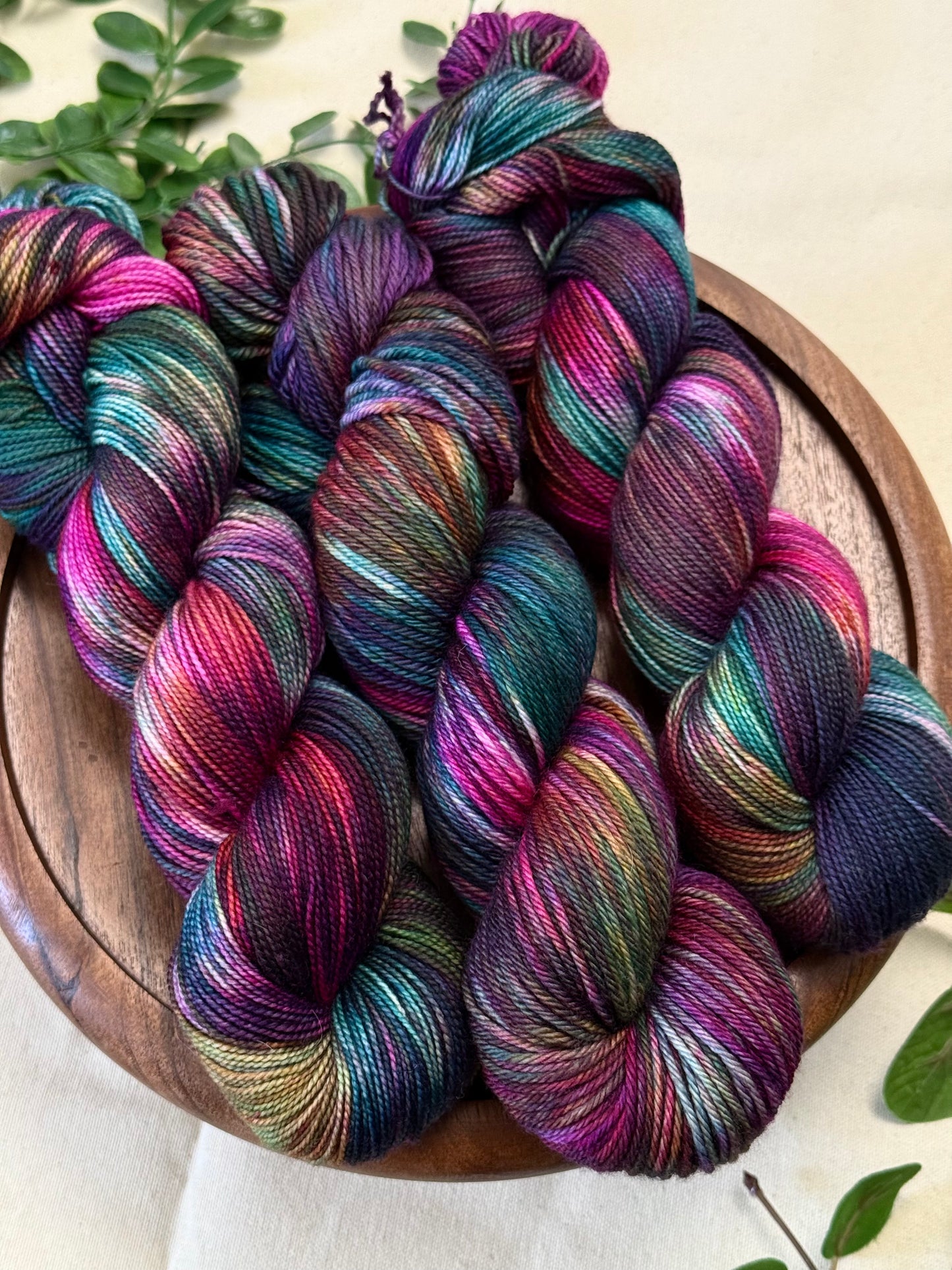 Several skeins of variegated yarn in vivid purple, teal, pink, blue, and olive tones arranged on a round wooden tray with small green leaves around the edges.