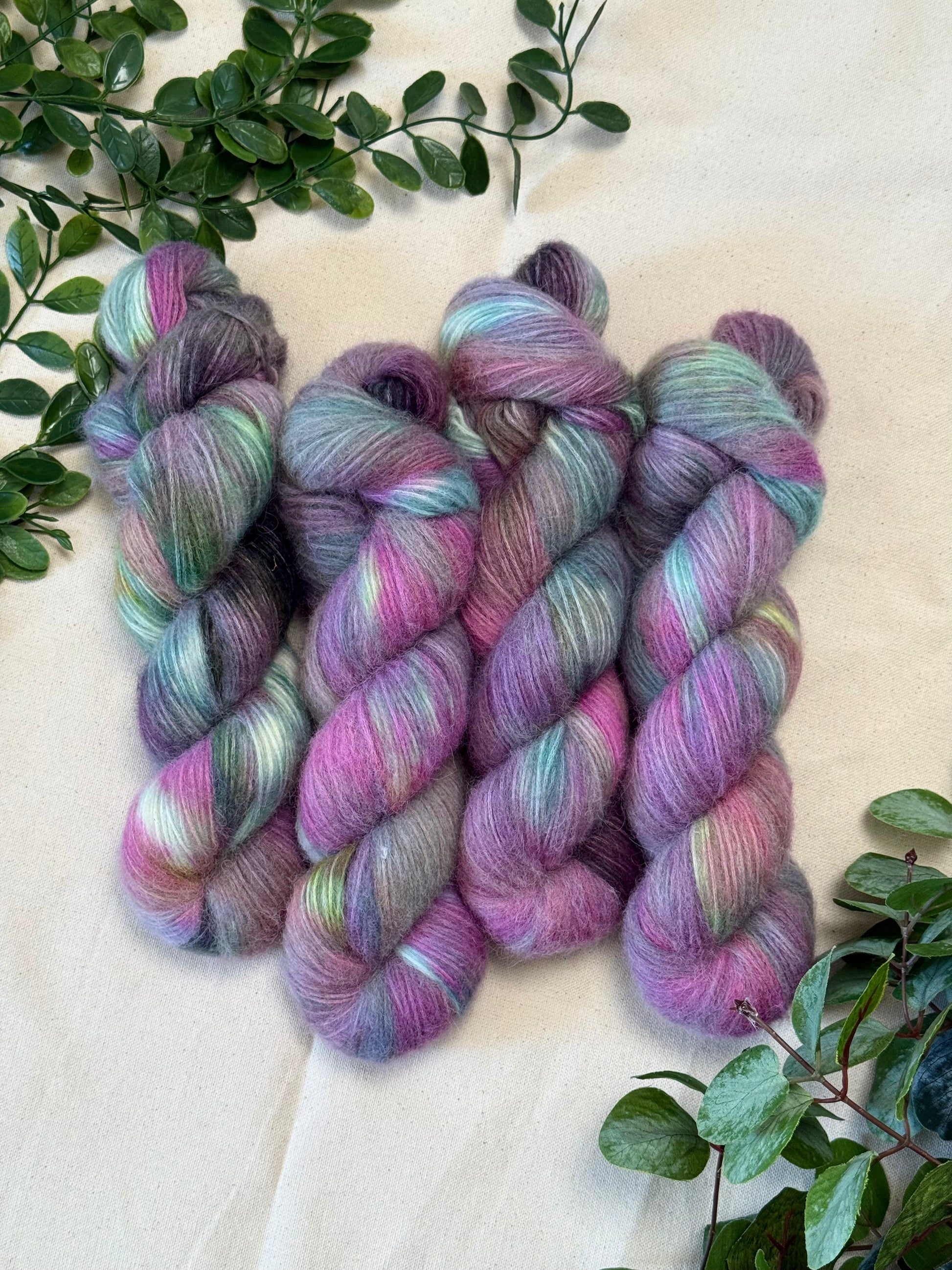 Four hanks of variegated merino wool yarn in shades of purple, pink, mint green, and blue, arranged on a beige fabric background with green foliage accents.
