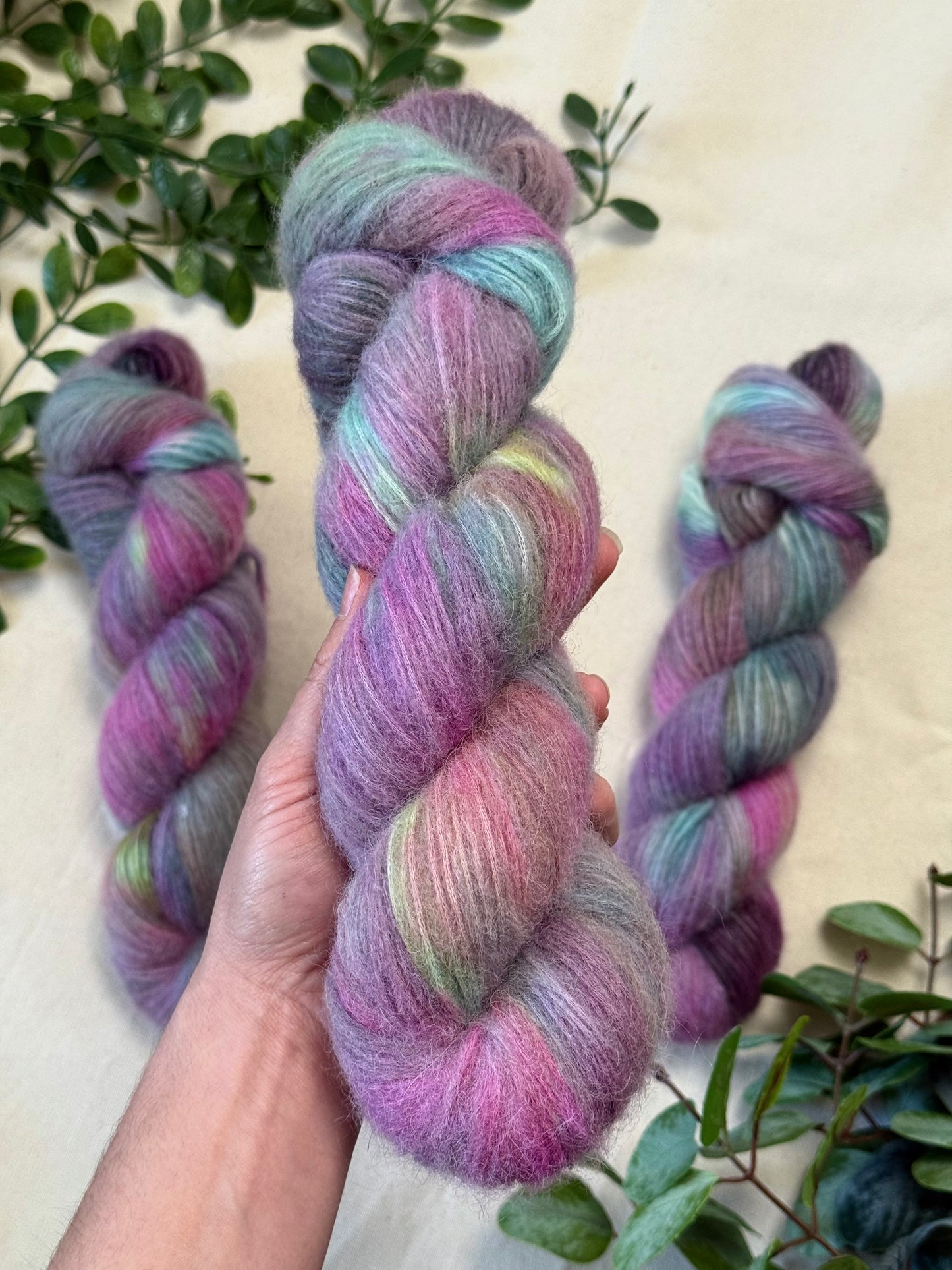 A hand holds a twisted skein of soft variegated yarn with pink, purple, blue, and mint tones, with two more skeins and green leaves arranged on a light beige surface.