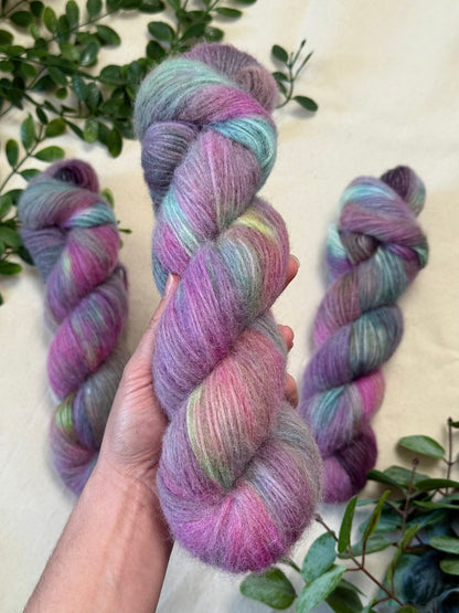 A hand holds a twisted skein of soft variegated yarn with pink, purple, blue, and mint tones, with two more skeins and green leaves arranged on a light beige surface.