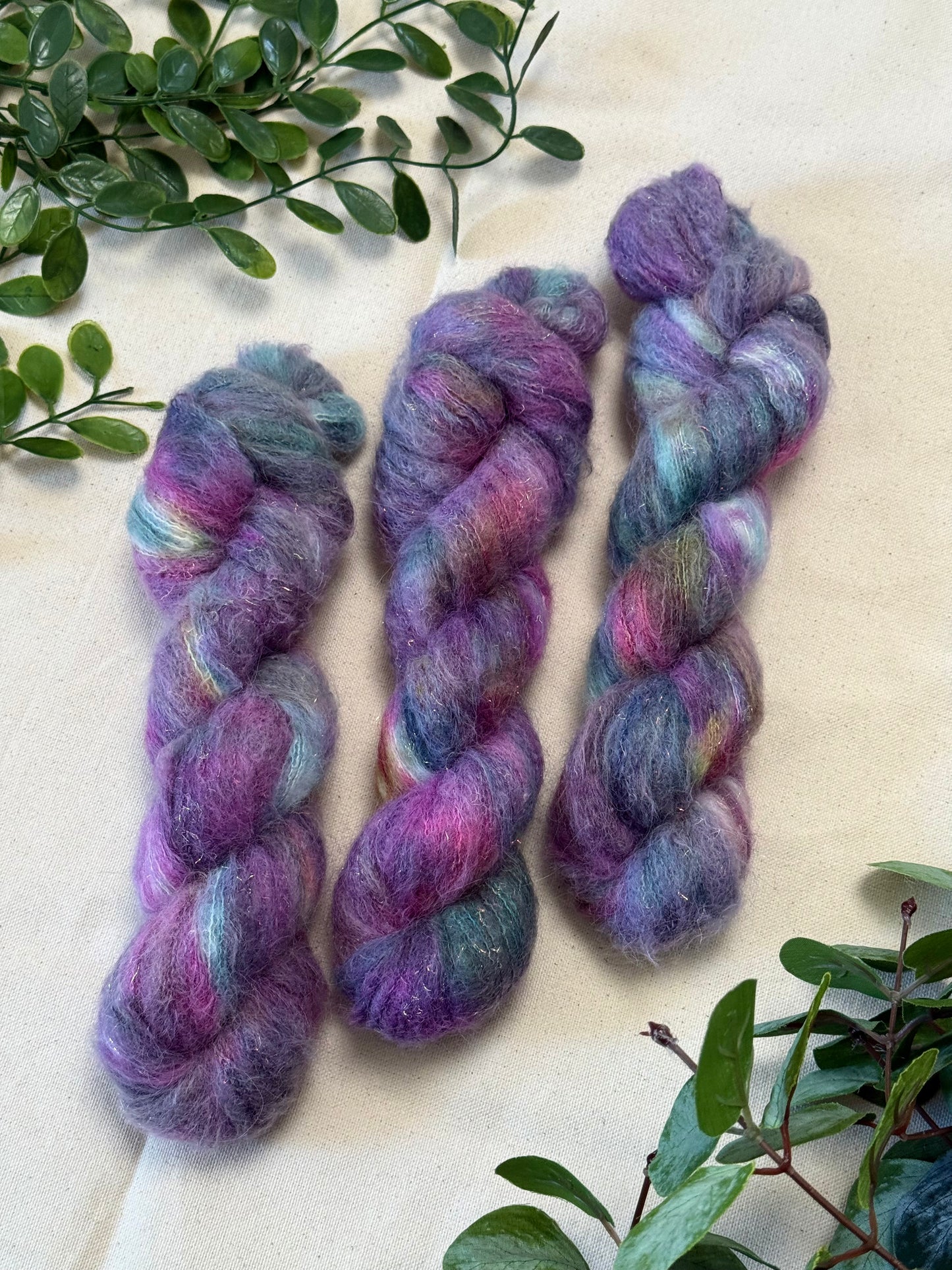 Three skeins of fluffy variegated yarn in shades of purple, pink, blue, and teal, arranged on a light beige fabric background with green leafy branches in the corners.