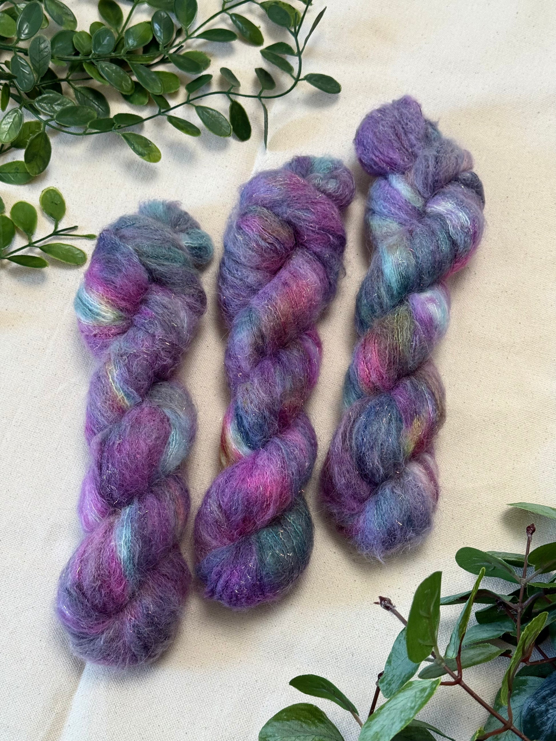 Three skeins of fluffy variegated yarn in shades of purple, pink, blue, and teal, arranged on a light beige fabric background with green leafy branches in the corners.