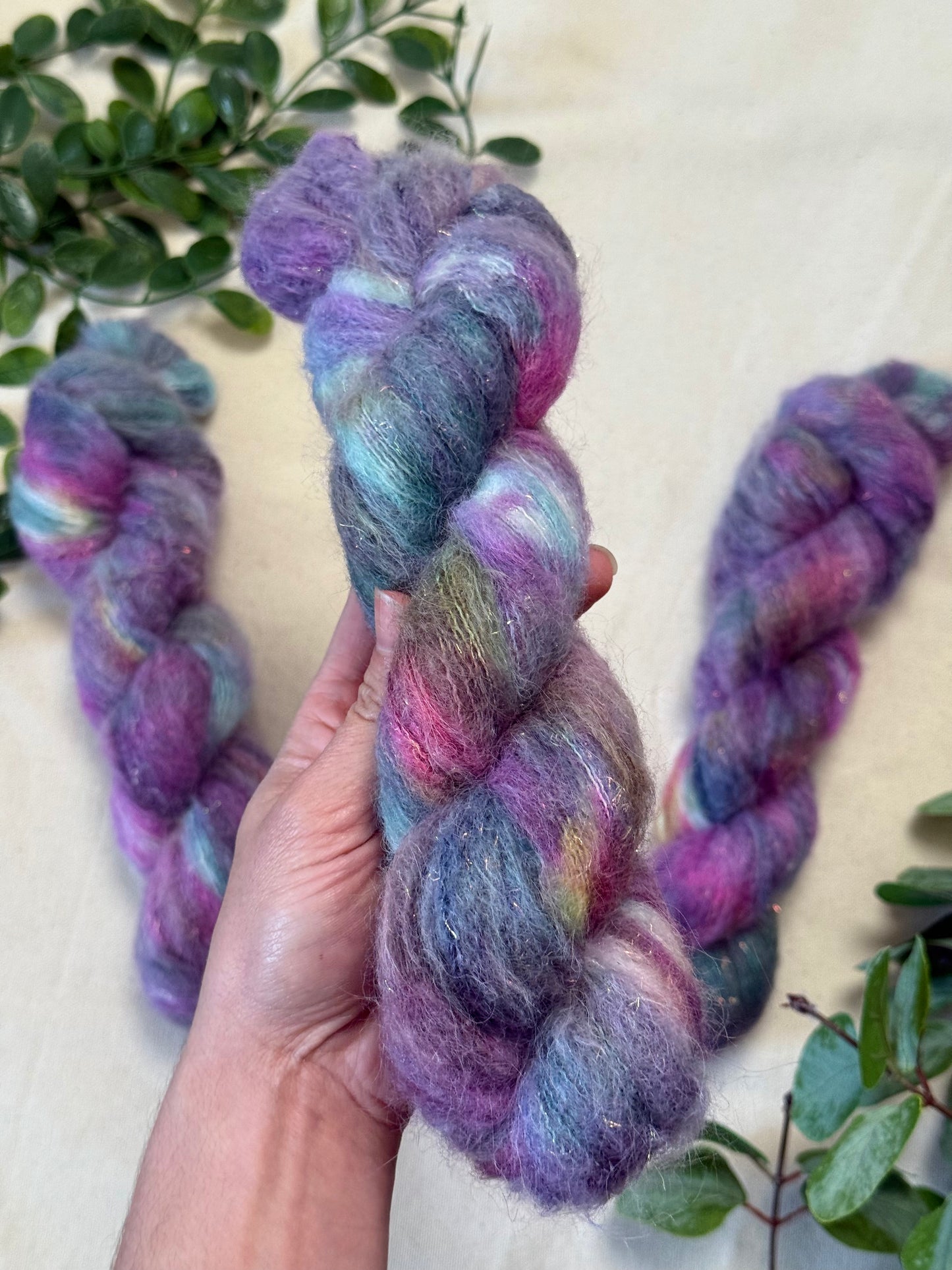 A hand holds a twisted skein of fluffy variegated yarn with purple, pink, blue, and green tones, with two more skeins and green leaves in the background.