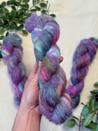 A hand holds a twisted skein of fluffy variegated yarn with purple, pink, blue, and green tones, with two more skeins and green leaves in the background.