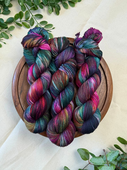 Three hanks of variegated yarn in teal, purple, pink, and green hues, arranged on a round wooden tray over a cream fabric background with green leaves in the corners.