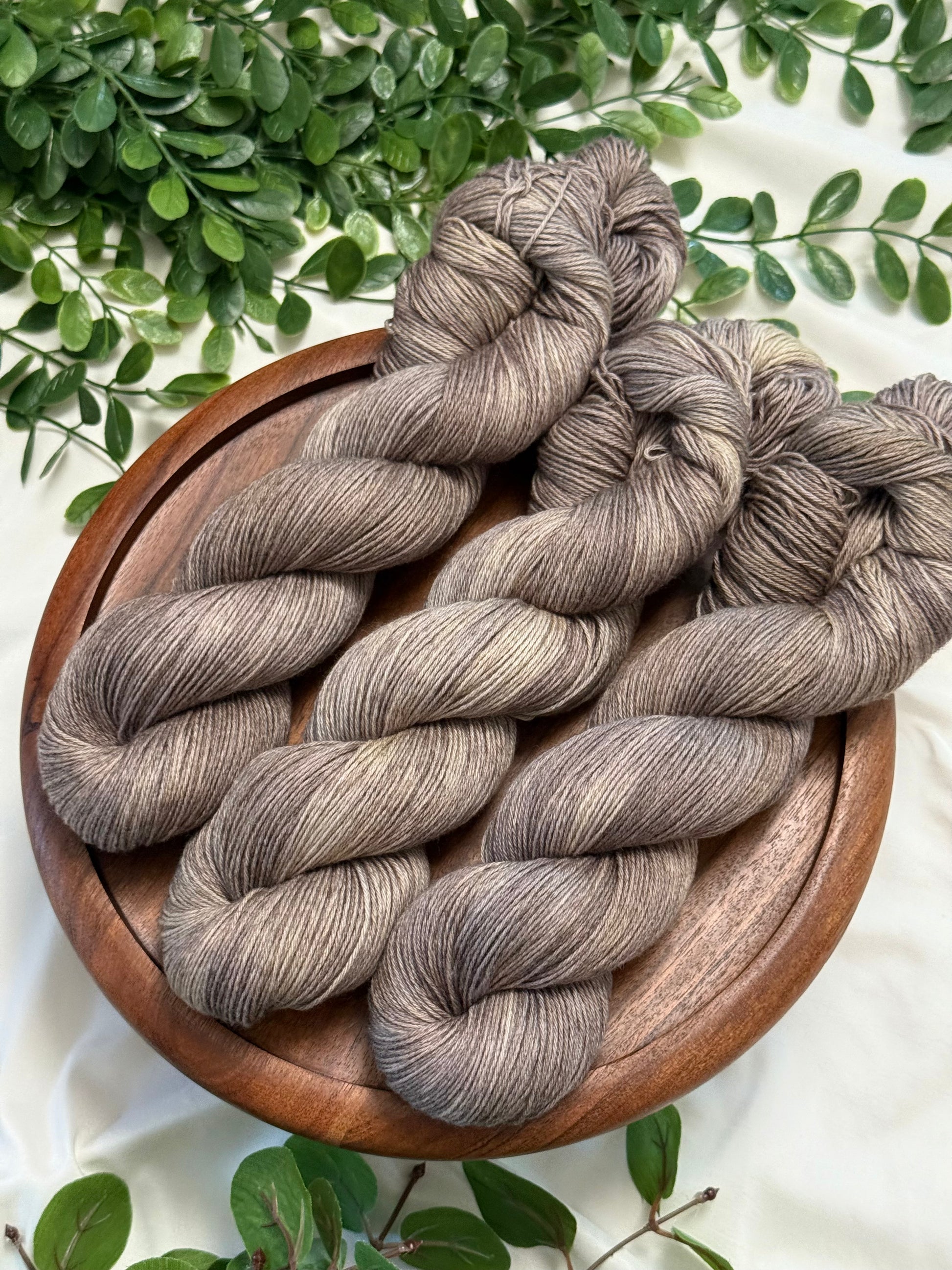 Three skeins of variegated gray and beige yarn arranged on a round wooden plate, with green foliage around the edges on a white fabric background.
