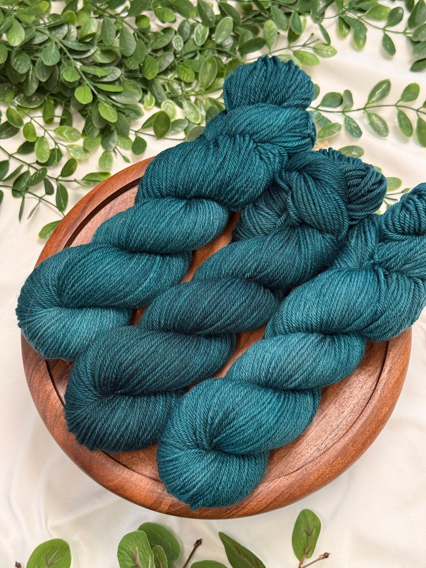 Three skeins of teal blue yarn arranged on a round wooden plate, with green leaves around it on a light fabric background.