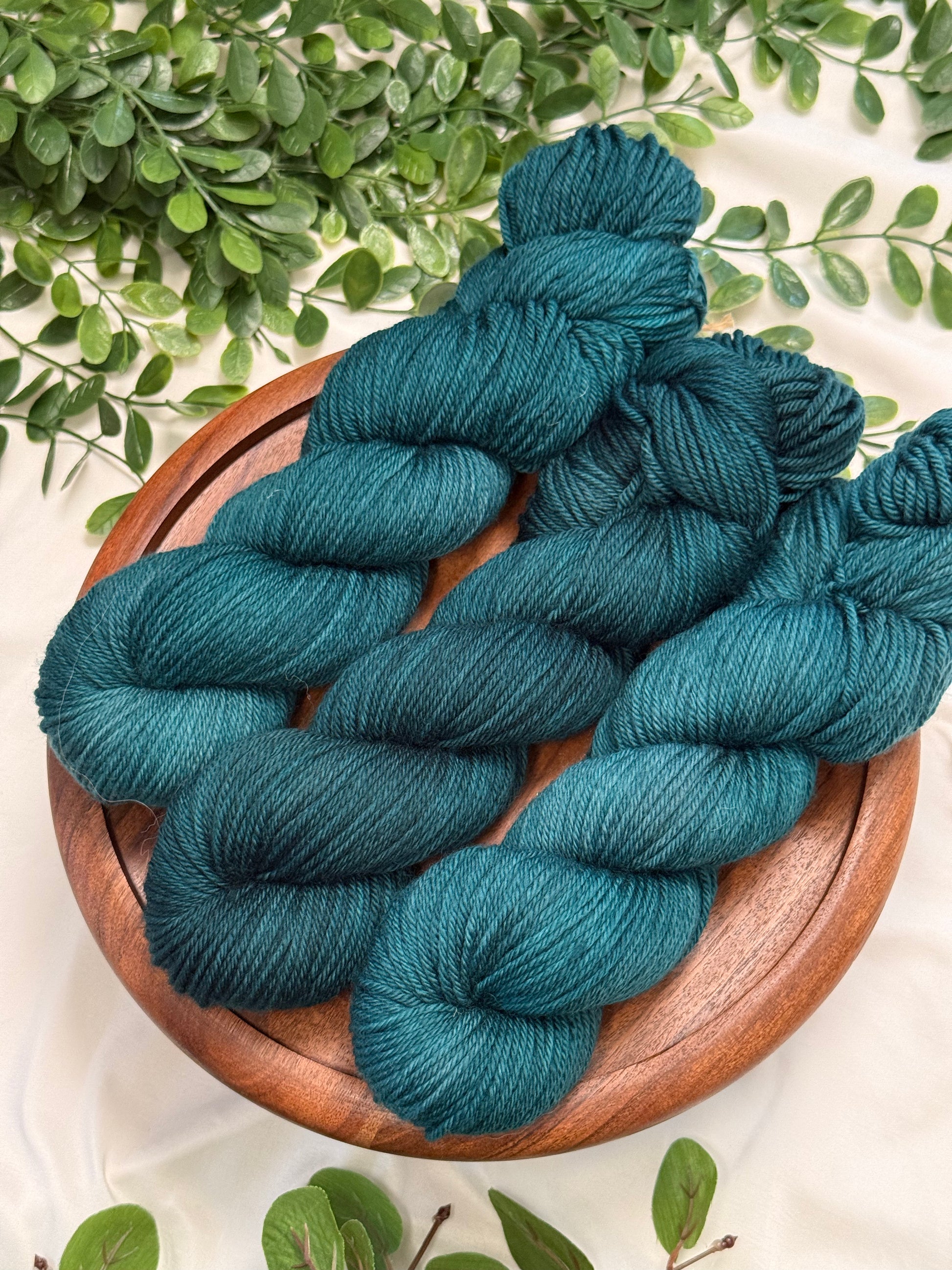 Three skeins of teal blue yarn arranged on a round wooden plate, with green leaves around it on a light fabric background.