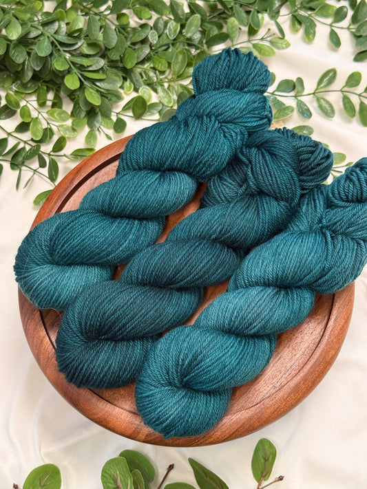Three skeins of teal blue yarn arranged on a round wooden plate, with green leaves around it on a light fabric background.