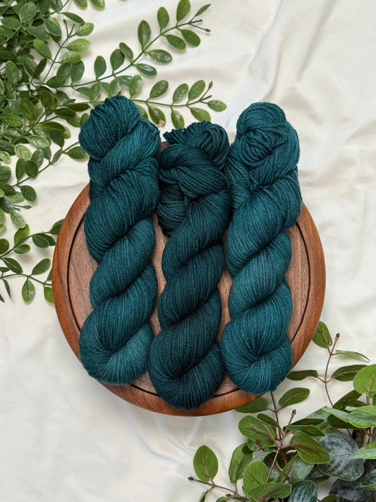 Three skeins of dark teal yarn arranged on a round wooden tray, surrounded by green leaves on a white fabric background.