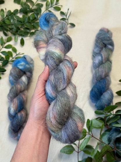 A hand holds a twisted skein of variegated wool yarn with blended blue, gray, lavender, and peach tones, with two more skeins and green leaves arranged on a light fabric background.