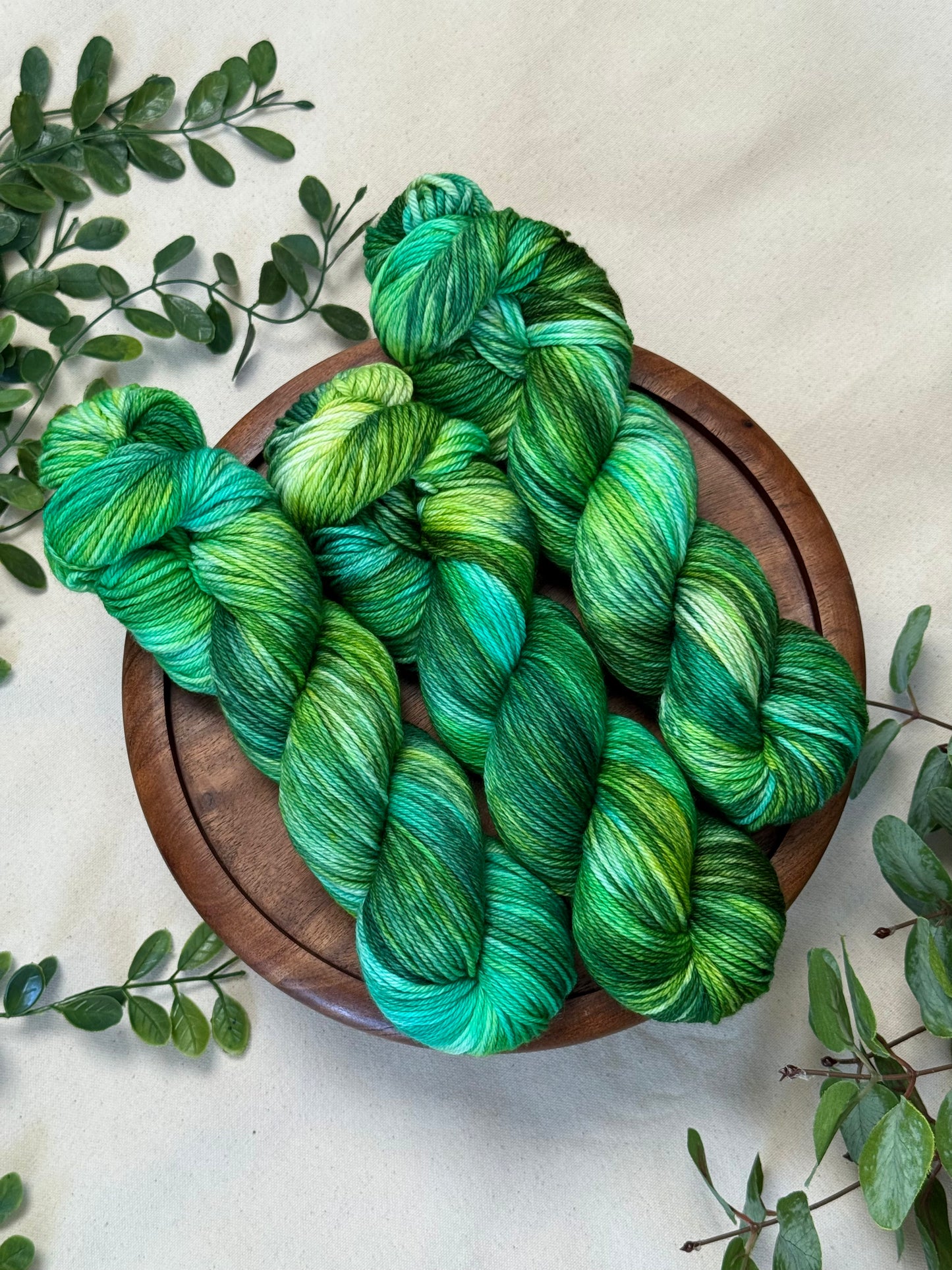 Three skeins of variegated green and blue yarn arranged on a round wooden tray, surrounded by green leaves on a light beige fabric background.