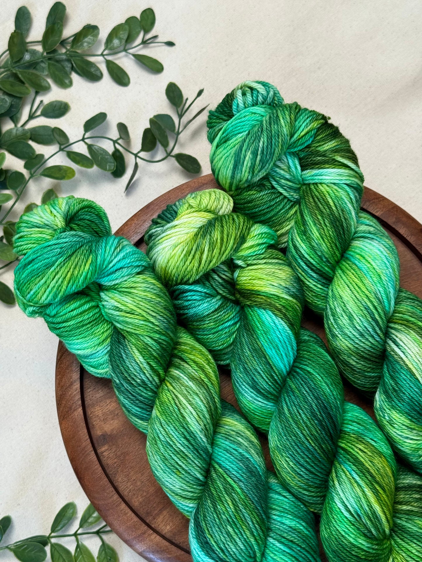 Three hanks of variegated green and teal yarn arranged on a round wooden tray, with small green leaves placed around it on a light neutral background.