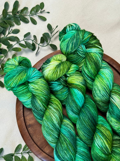 Three hanks of variegated green and teal yarn arranged on a round wooden tray, with small green leaves placed around it on a light neutral background.