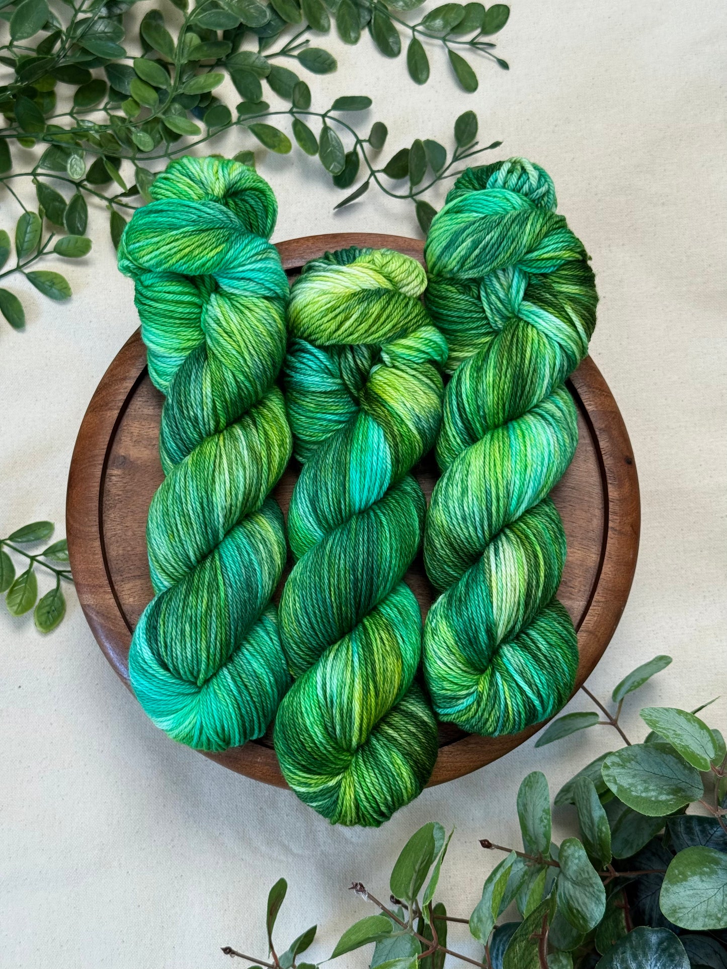 Three skeins of variegated green and teal yarn arranged on a round wooden tray, surrounded by green leaves on a light beige fabric background.