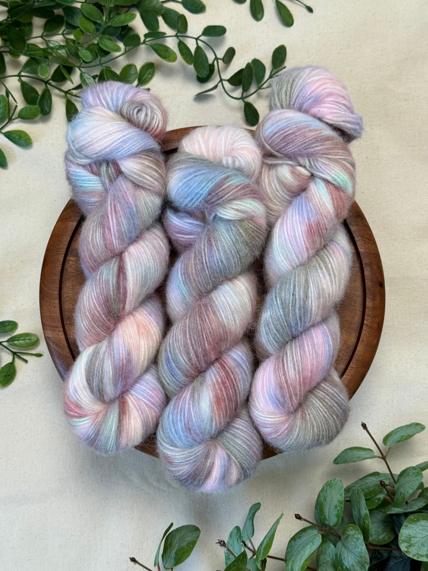 Three twisted hanks of variegated yarn in pastel pink, lavender, and pale blue tones, arranged on a round wooden tray with green leaves around it on a light fabric background.