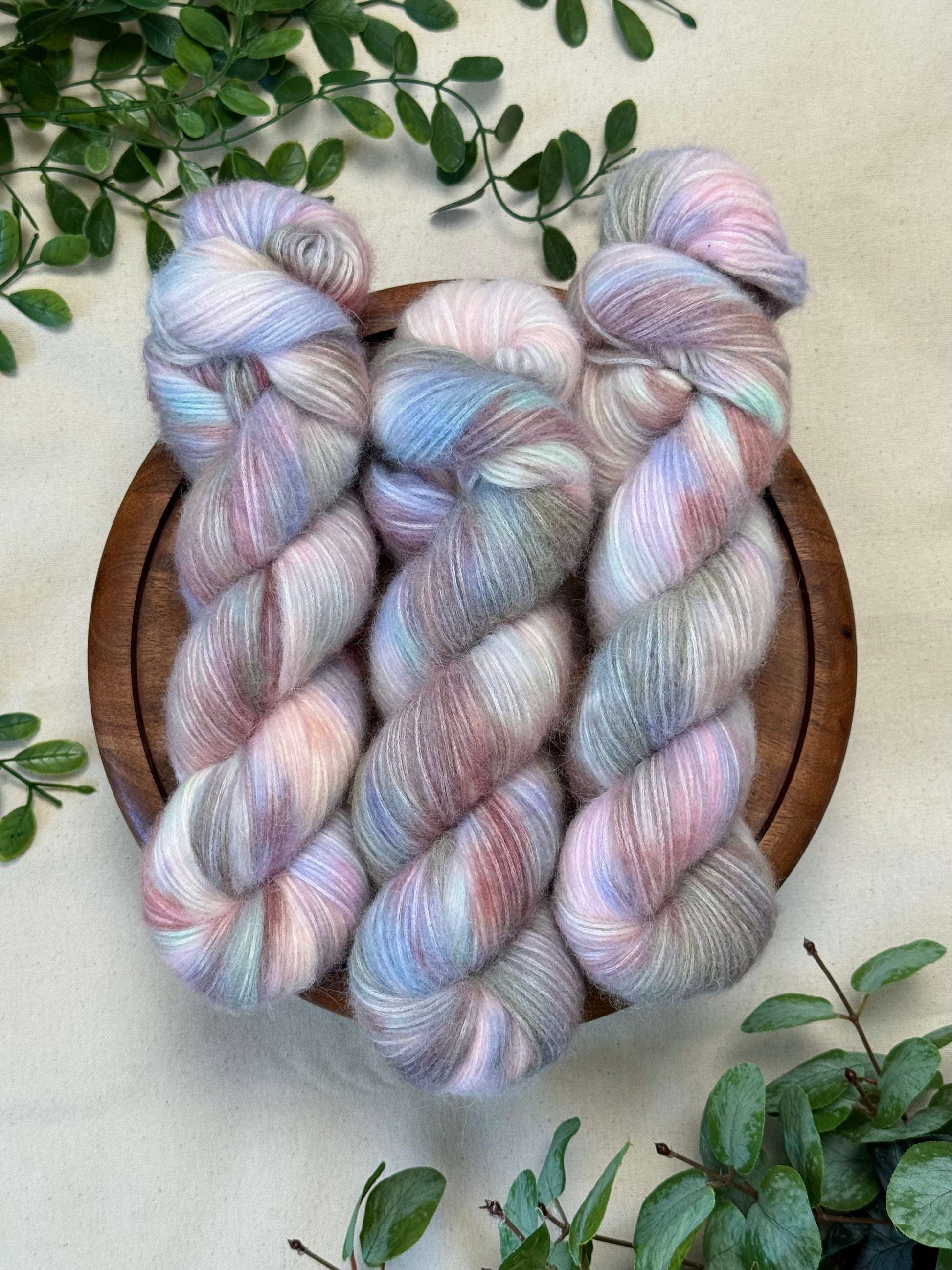 Three twisted hanks of variegated yarn in pastel pink, lavender, and pale blue tones, arranged on a round wooden tray with green leaves around it on a light fabric background.