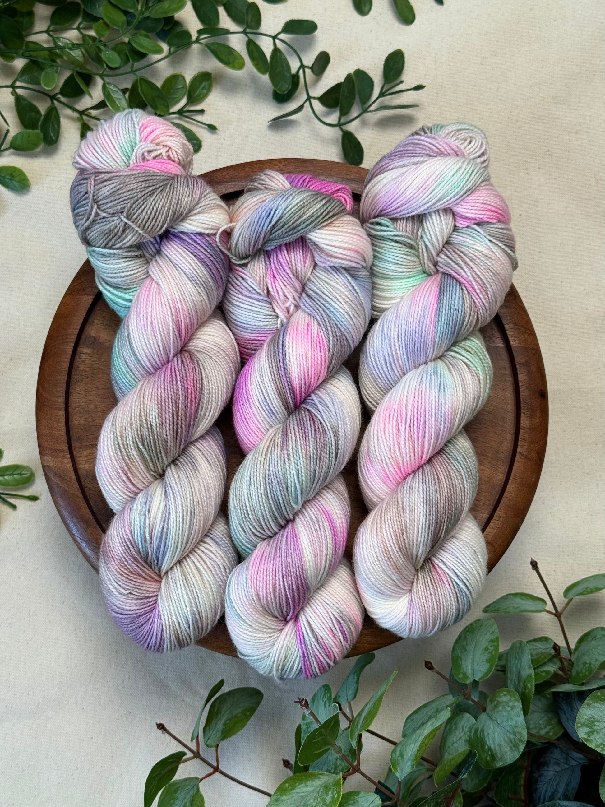 Three hanks of variegated yarn in pastel pink, lavender, mint, and gray tones are arranged on a round wooden tray, surrounded by green leaves on a light neutral background.