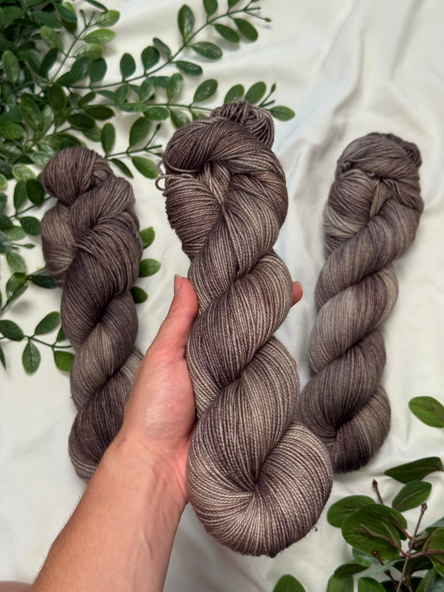 Three hanks of variegated yarn in a taupe-brown and gray colorway are displayed on a white fabric background with green leaves. A hand is holding the center hank to show its texture and thickness.
