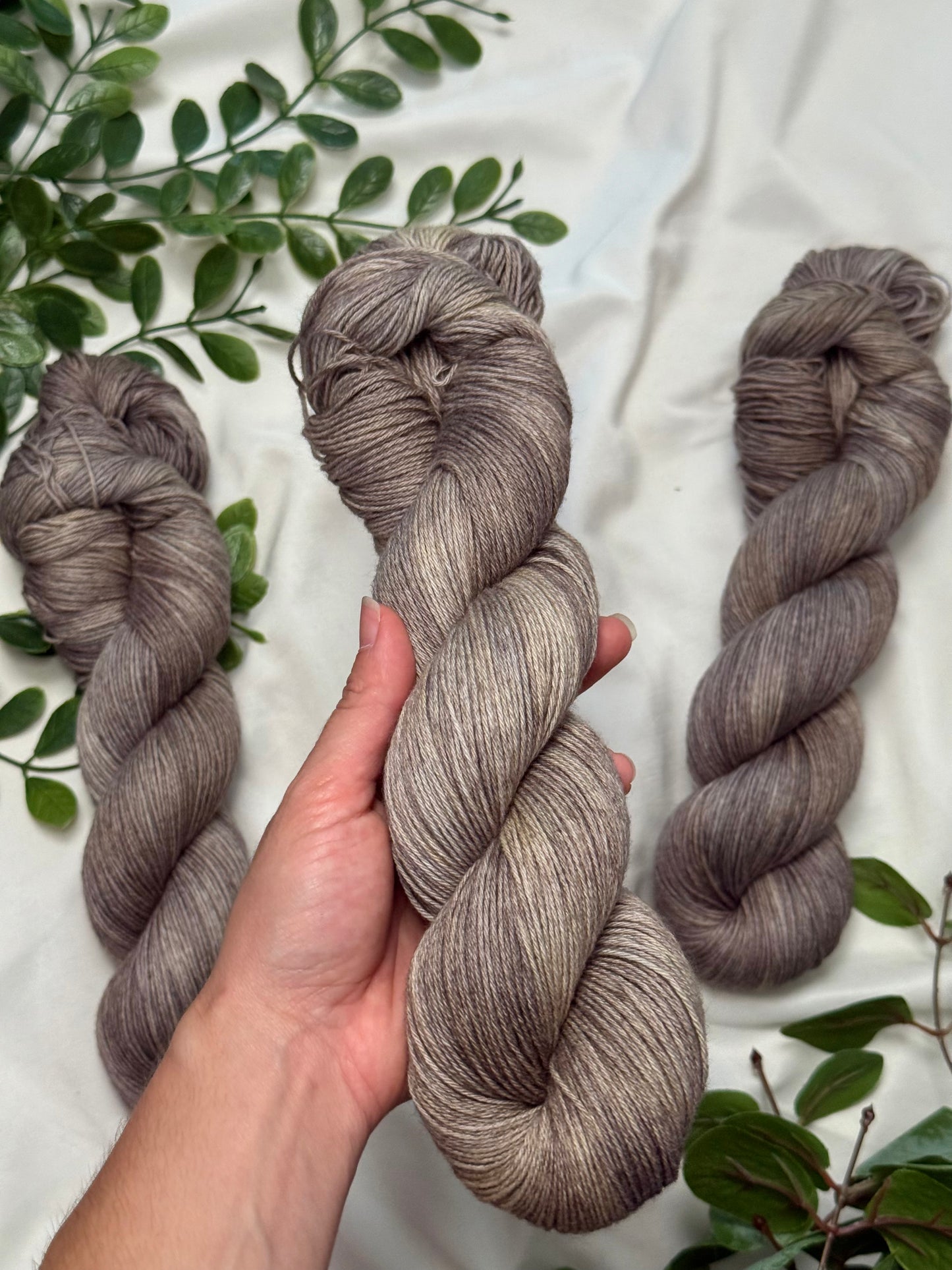 Three hanks of variegated gray-brown yarn are displayed on a white fabric background with green leaves. A hand is holding the center hank, showing the texture and color variation of the yarn.