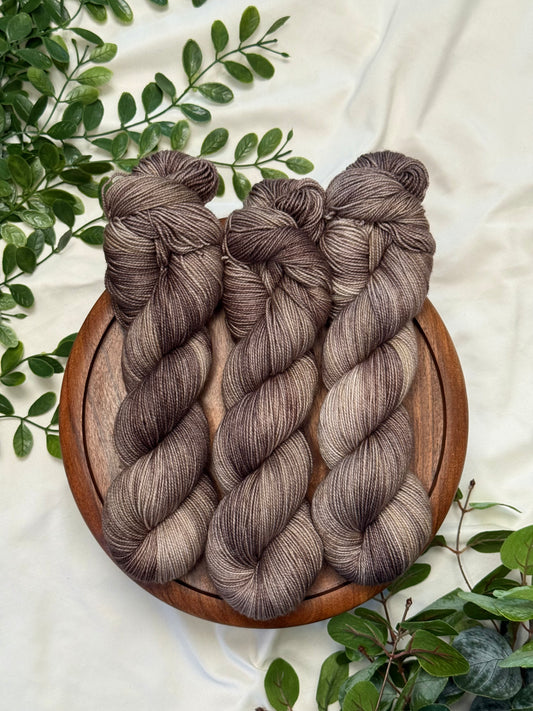 Three hanks of variegated yarn in earthy taupe-brown and gray tones, arranged on a round wooden tray with green leaves on a cream fabric background.