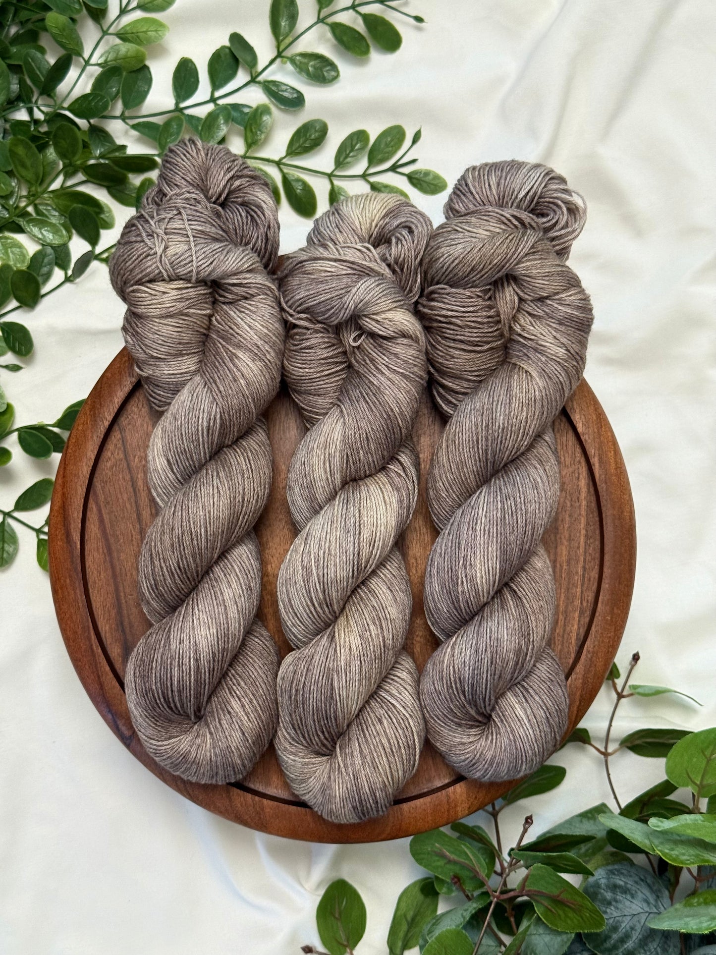 Three twisted hanks of heathered taupe-gray yarn arranged on a round wooden board, surrounded by green leaves on a white fabric background.